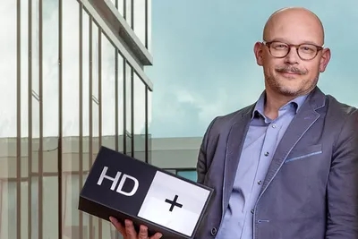 Ein Mann im Anzug hält eine „HD+“-Box in der Hand und steht vor einem modernen Glasgebäude mit bewölktem Himmel im Hintergrund.