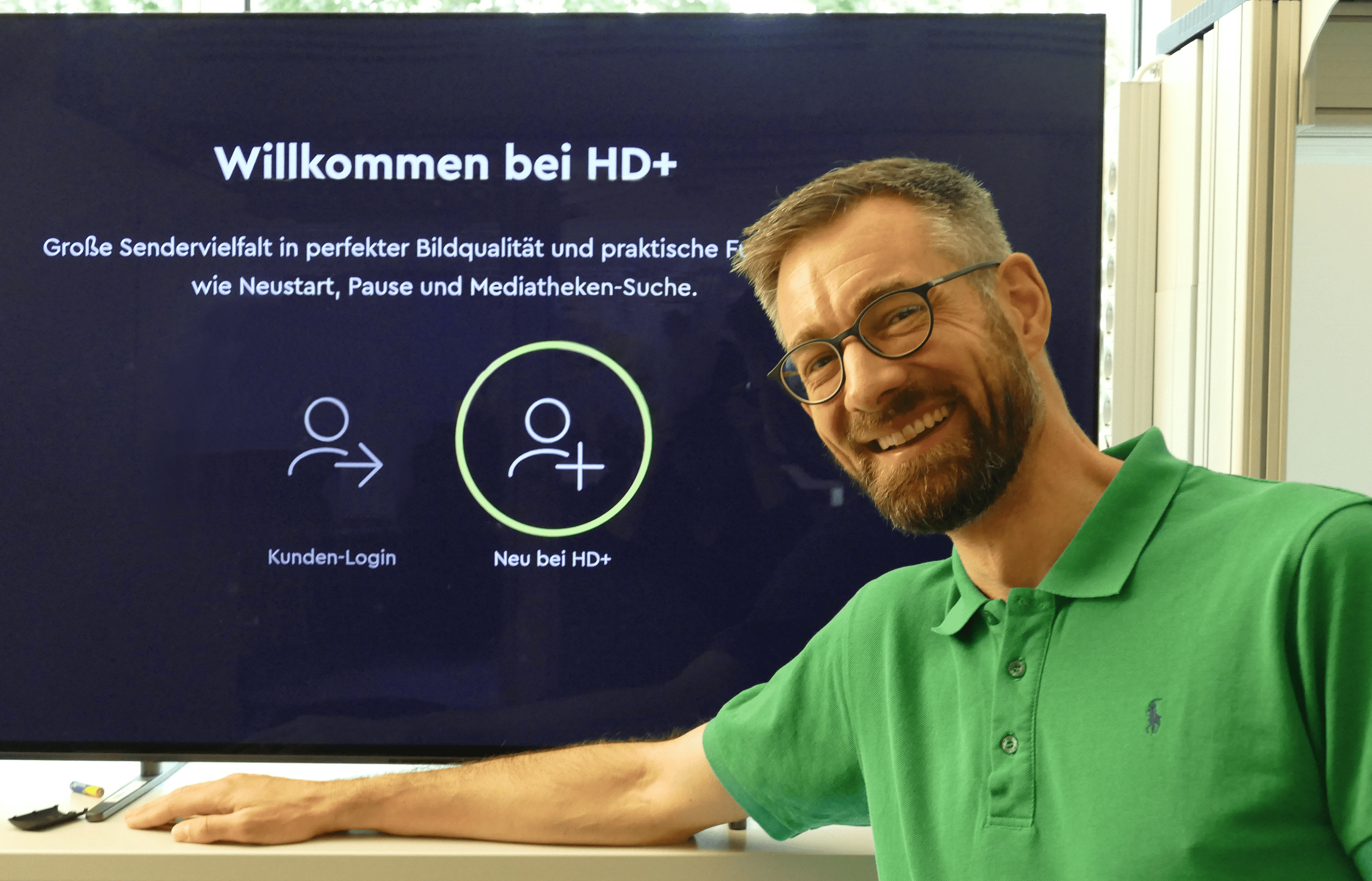 Lächelnder Mann in einem grünen Polohemd, der neben einem Bildschirm steht, auf dem „Willkommen bei HD+“ mit Anmeldeoptionen angezeigt wird.