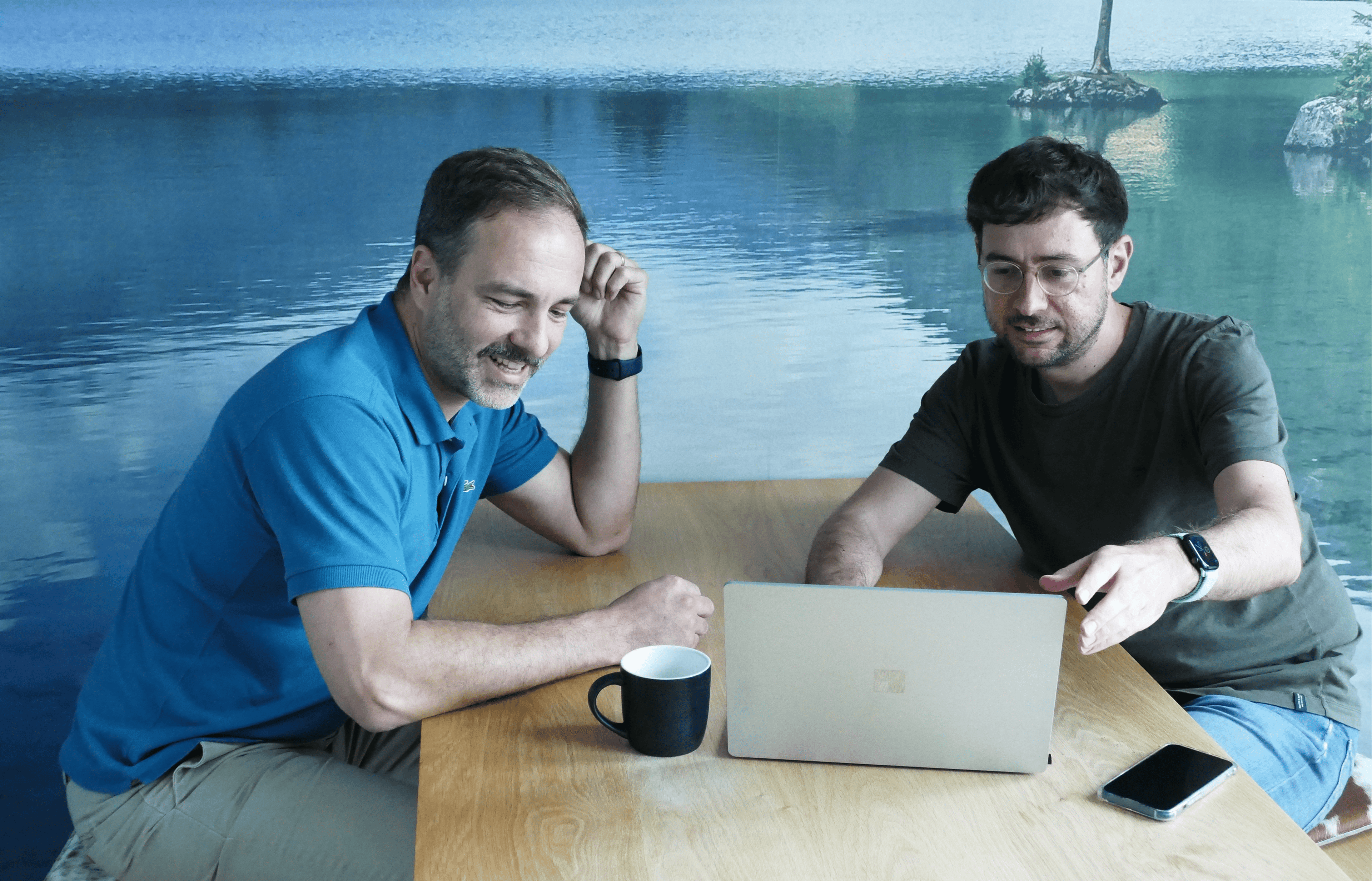Zwei Männer sitzen an einem Tisch, lächeln und schauen auf einen Laptop. Einer zeigt auf den Bildschirm. Auf dem Tisch stehen eine Tasse und ein Telefon. Im Hintergrund ist ein See zu sehen.