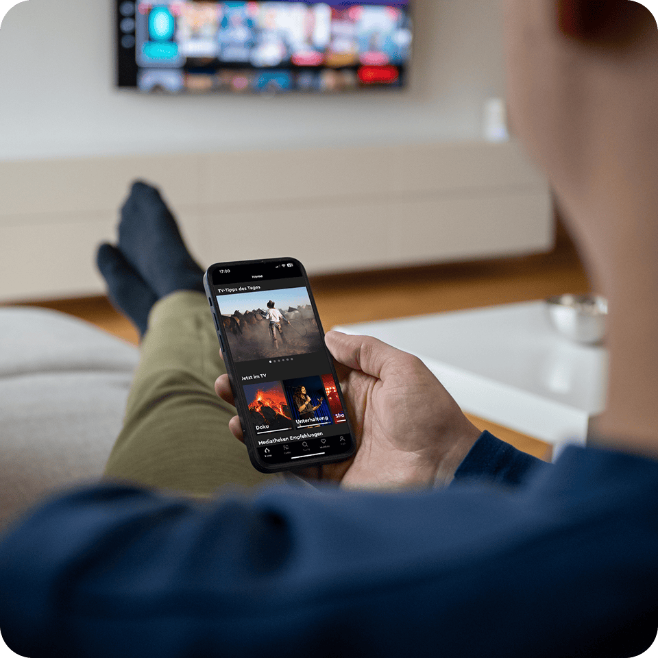 Person in blauem Hemd, die mit einem Smartphone auf einer Couch sitzend eine Streaming-App nutzt, im Hintergrund ist ein Fernseher zu sehen.