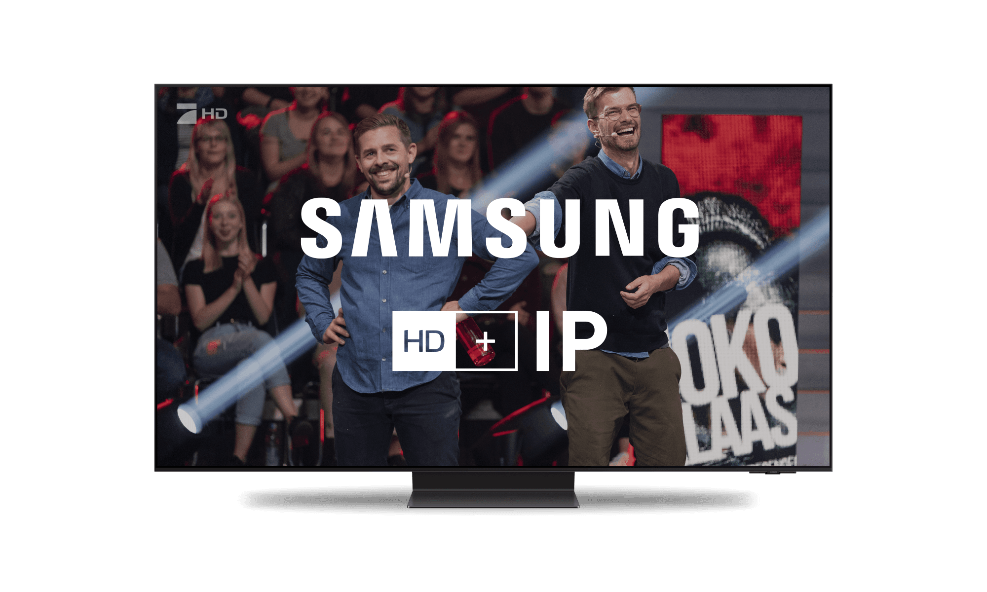 Zwei lächelnde Männer auf einer Fernsehshow-Bühne mit sitzendem Publikum; Samsung-HD-Logo auf dem Bildschirm angezeigt.